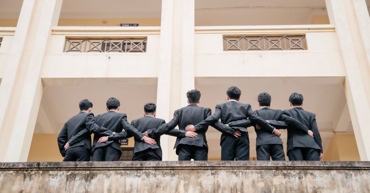Seven men in suits standing with arms around each other, viewed from behind in an outdoor setting.